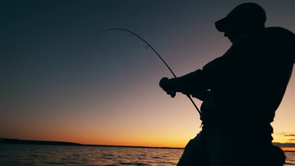 En man rullar upp en fiskespö i en solnedgång sjö — Stockvideo