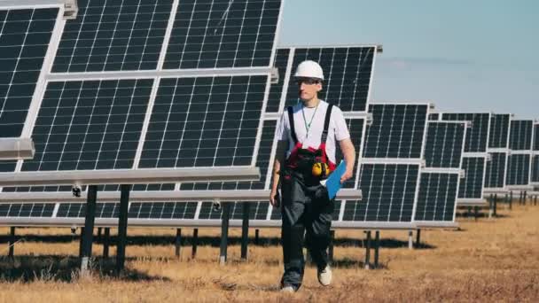 Güneş enerjisi santrali yakınlarında alternatif enerji işçisi. Erkek teknisyen sahada güneş pilleriyle yürüyor. — Stok video