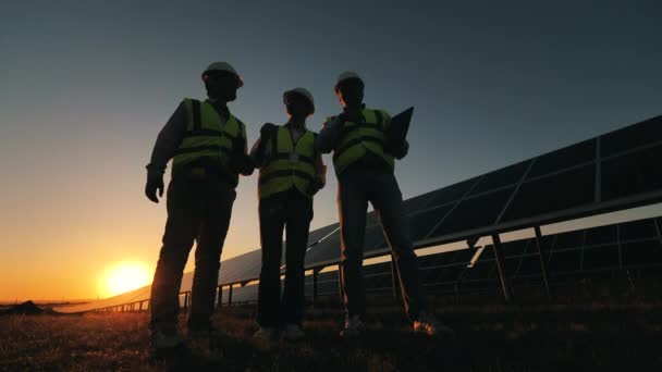 Teamet av ingenjörer diskuterar ett projekt nära solbatterier under solnedgången. Alternativa energiarbetare nära solkraftverk. — Stockvideo