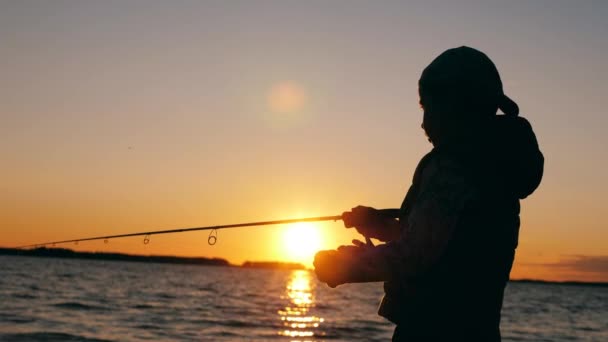 Um menino está lidando com um pólo de pesca perto de um lago pôr do sol — Vídeo de Stock