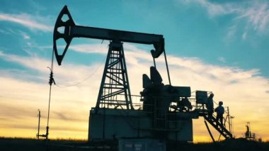 Petrol pompalama ünitesi ve gün batımında onunla çalışan iki teknisyen. Petrol kuyusu, ham petrol, sondaj kulesi, petrol sahası, ham petrol fiyatı, petrol ve doğalgaz, petrol varili, petrol endüstrisi konsepti.