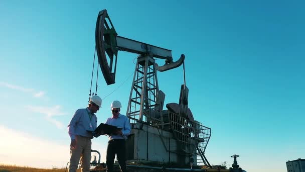 Poço de petróleo, petróleo bruto, equipamento de perfuração, campo de petróleo, preço do petróleo bruto, petróleo e gás, barril de petróleo, conceito da indústria do petróleo. Dois engenheiros estão falando ao lado de uma unidade de bombeamento de óleo — Vídeo de Stock