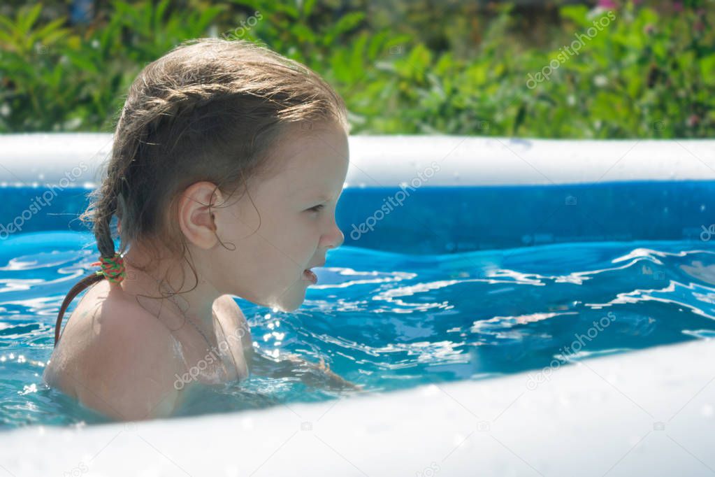The girl smiles, squints from the sun, the child is pleased with the bathing and swimming in the water in the inflatable pool. Summer vacation for families