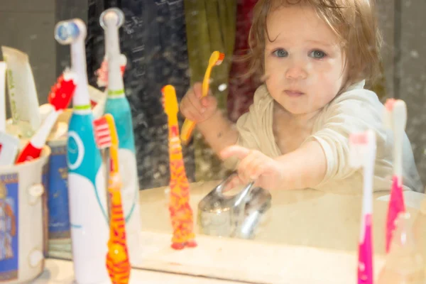 透过一个肮脏的牙膏飞溅的镜子看一个小女孩 孩子刷牙 从浴室里的第一颗乳牙上清除牙菌斑 浴室里的一切都很混乱 — 图库照片