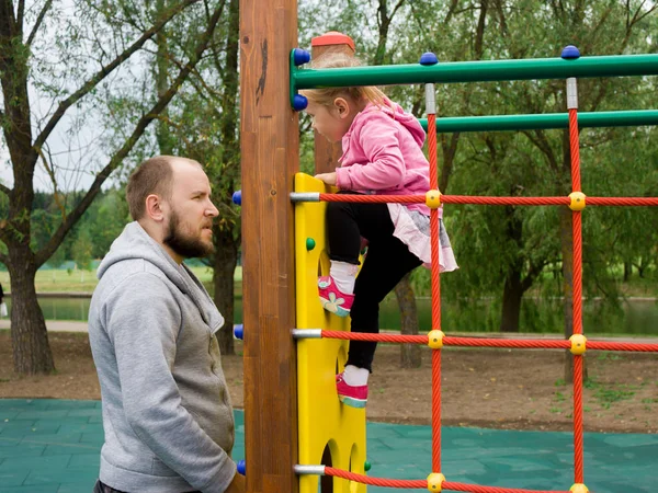 Babası merdiven ve yerel park crossbars tırmanabilir miyim onun küçük bebek kızı, güvenli oyun üzerinde yatay çubuk yanında duruyor.