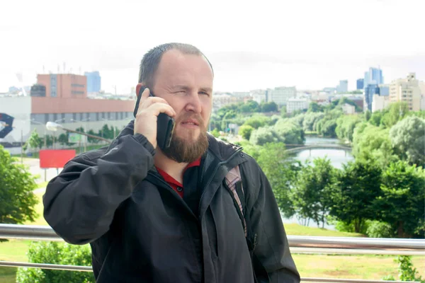 Business mature male tourist with a beard talking on the phone and make an appointment on the background of the spring cityscape.