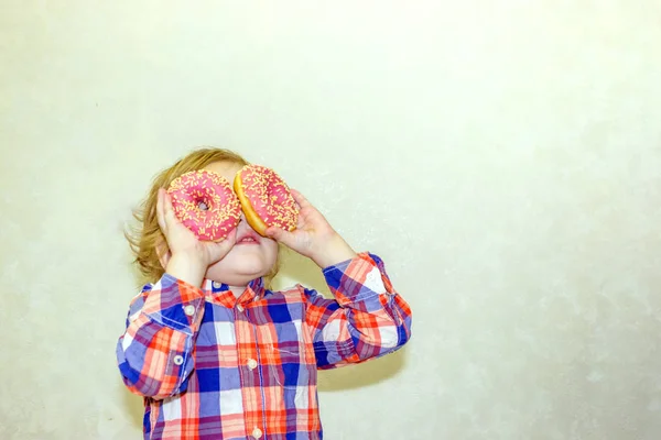 一个小孩在吃饭前涉足和两个新鲜的甜甜圈玩耍 一个孩子把甜甜圈放在眼睛附近 透过眼镜看洞 — 图库照片