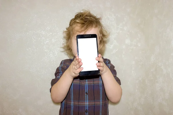 The little boy is hiding behind the phone. The child shows a blank white phone screen. Kid looks out from behind the smartphone with interest.