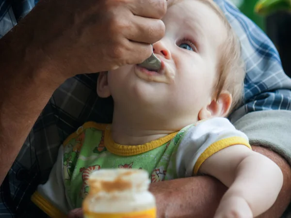 一个小女孩坐在她祖父的怀里 用勺子吃水果泥 在儿童中引入补充食物 大粗糙的手轻轻地支持婴儿 用勺子喂食 — 图库照片