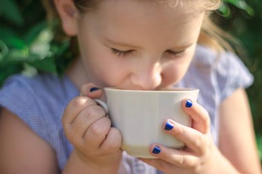 Mutlu küçük kız yeşil arka planda beyaz fincandan aromatik çiçek çayı tadıyor.