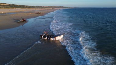 Costa da Caparica, Portekiz 22 Ağustos 2025: Balıkçıların teknelerini okyanus dalgalarına sürdükleri hava manzarası. Sahilde geleneksel balıkçılık sahnesi, takım çalışması ve deniz yaşamı yukarıdan ele geçirildi.