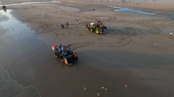 Costa da Caparica, Portekiz 22 Ağustos 2025: Balıkçılar gün batımında okyanustan ağlar, plajda traktörler ve botlar çekiyor, insanlar avı izliyor, martılar yukarıda uçuyor. Yerel balıkçılık endüstrisi. Havadan