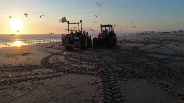 Costa da Caparica, Portekiz 22 Ağustos 2025: Balıkçılar gün batımında okyanustan ağlar, plajda traktörler ve botlar çekiyor, insanlar avı izliyor, martılar yukarıda uçuyor. Yerel balıkçılık endüstrisi. Havadan