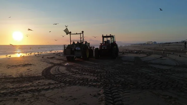 Costa da Caparica, Portekiz 22 Ağustos 2025: Balıkçılar gün batımında okyanustan ağlar, plajda traktörler ve botlar çekiyor, insanlar avı izliyor, martılar yukarıda uçuyor. Yerel balıkçılık endüstrisi. Havadan