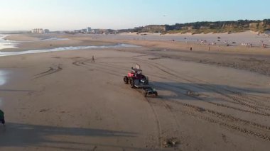 Costa da Caparica, Portekiz 22 Ağustos 2025: Balıkçılar gün batımında okyanustan ağlar, plajda traktörler ve botlar çekiyor, insanlar avı izliyor, martılar yukarıda uçuyor. Yerel balıkçılık endüstrisi. Havadan