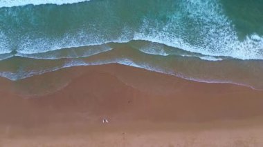 Üzerinde kumlu bir sahil manzarası olan mavi turkuaz dalgalar kıyıya vuruyor, sahil boyunca yürüyen sörfçüler, gün batımından önce okyanus..