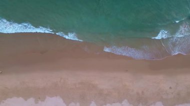 Üzerinde kumlu bir sahil manzarası olan mavi turkuaz dalgalar kıyıya vuruyor, sahil boyunca yürüyen sörfçüler, gün batımından önce okyanus..