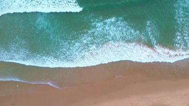 Üzerinde kumlu bir sahil manzarası olan mavi turkuaz dalgalar kıyıya vuruyor, sahil boyunca yürüyen sörfçüler, gün batımından önce okyanus..