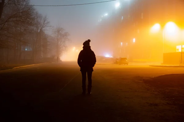 Egy ember sötét sziluettje áll a ködben a házak és fák fényeinek hátterében. Vízszintes tájolás. — Stock Fotó