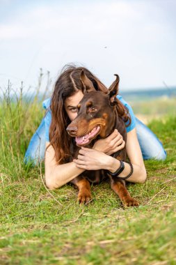 Kahverengi ve bronzlaşmış doberman köpeği olan uzun saçlı bir kız güneşli bir günde yeşil çimlerin üzerinde yatıyor. Arka planda bulanık bir portre. Dikey oryantasyon. İzleyiciye bir bak..