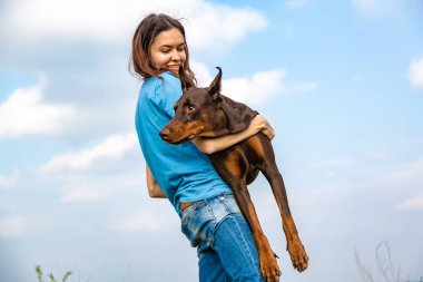Mavi tişörtlü ve kotlu bir kız kahverengi ve esmer bir Doberman Dobermann 'ı bulutlu gökyüzünün ellerinde tutuyor. Yatay yönelim. 