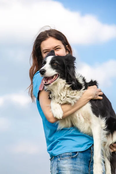 A girl holds a black and white border collie dog in her hands and hugs and smiling. Portrait against a bright blue sky with clouds. Vertical orientation. — Stock Photo, Image