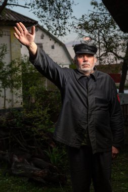 Eski moda siyah giysileri ve kırsal kesimin arka planında kaldırılmış deri şapkası olan ciddi yaşlı bir adam. Leninist jest. Kameraya bakıyorum. Dikey yönelim. 