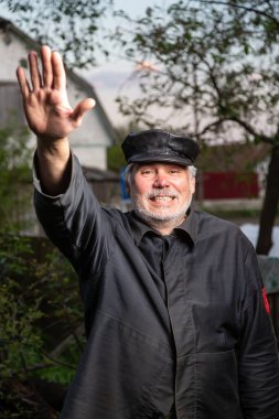 Eski moda siyah giysileri ve kırsal kesimin arka planında kaldırılmış deri şapkası olan neşeli yaşlı bir adam. Leninist jest. Kameraya bakıyorum. Dikey yönelim. 