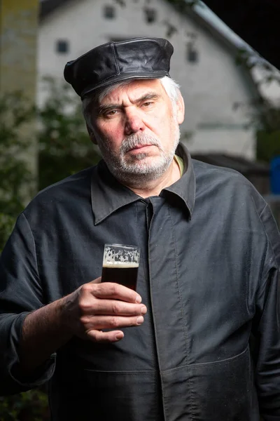 Serious kind sad elderly man with black old-fashioned old clothes and a leather cap with a glass of beer in his hand. Looks at the camera. Vertical orientation.