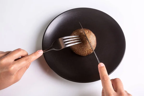 En person håller en bestickgaffel och kniv och skär en rå tröja ko svamp på en svart tallrik på en vit bakgrund. Händerna i ramen. Horisontell inriktning. Högkvalitativt foto — Stockfoto