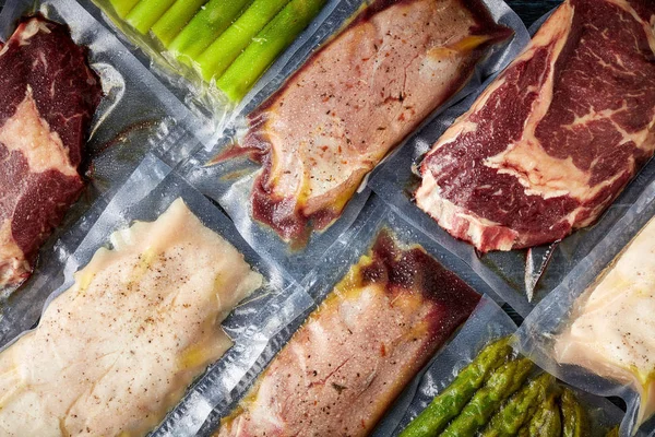 Duck breast, chicken breast, beef steak and asparagus vacuum sealed ready for sous vide cooking, on black background, top view