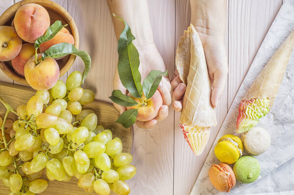 Руки молодой женщины с тортом и персиком, делающей выбор между ними. Женщина делает выбор между сладостями и фруктами. Здоровое и нездоровое питание. вид сверху, плоский
.