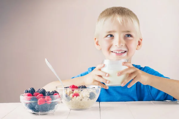 Happy little boy with milk mustache holding glass of milk or yogurt and eating porridge with berries. The concept of a healthy breakfast for children.