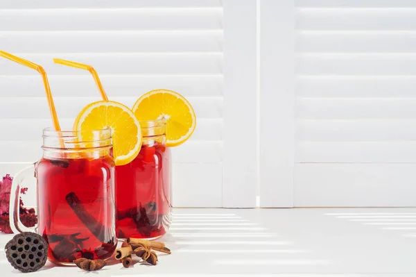 Two fruit tea in vintage glass jars with drinking straw against vintage white wooden shutters background. Vintage mason jars with ice red tea, cinnamon and star anise on white window sill
