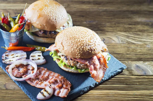 Fresh tasty burger with copy space. Close-up of two homemade burgers, sesame buns with succulent beef patties, grilled onions, bacon and fresh salad served on slate board on a rustic wood table.