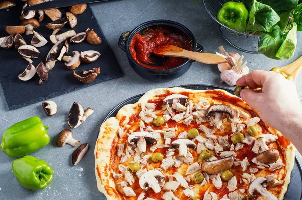 Pizza preparation, raw dough surrounded by ingredients. A man\'s hand spreads olives for pizza dough.