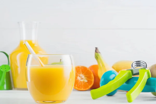 A glass of fresh orange juice and fruits, tangerine, apple, banana, dumbbell, hand gripper on white wooden background. Healthy lifestyle and diet concept. Healthy eating for weight loss.