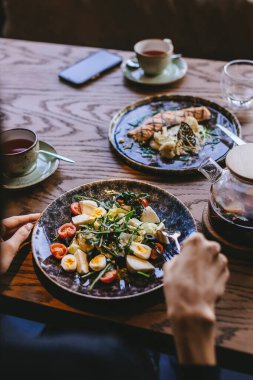 POV - Unrecognized lady sitting at wooden table having lunch and enjoying vibrant healthy vegetarian salad with fresh vegetables and eggs alongside grilled fish at modern restaurant.
