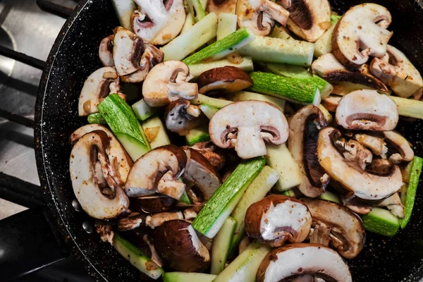 Champingon mushrooms and green zucchini cooking on a silver kitchen stove. Delicious vegetables vegan food sizzling on the black frying pan. Nutritious food fiber healthy diet. Restaurant homemade