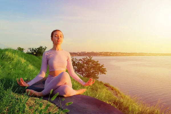 Hermosa mujer sana adulta joven meditando haciendo yoga en la colina verde junto al mar al aire libre. mujer en un traje rosa una vaca pose pilates. vida consciente, tratamiento de ansiedad cuidado de la salud mental . — Foto de Stock