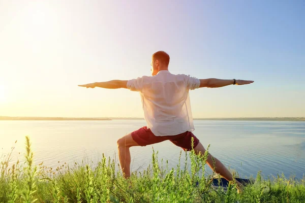 Fit muscular man doing yoga by the water on the grass with hands stretch. Orange sunlight sunset tranquility at lake. Male back in white office shirt. Pilates warrior pose Healthy spine workout
