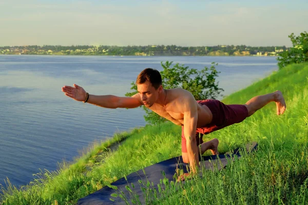 Fit muscular man doing yoga by the water on the grass with hands stretch. Orange sunlight tranquility at lake. Male back in white office shirt. Pilates warrior pose. Healthy spine fitness workout