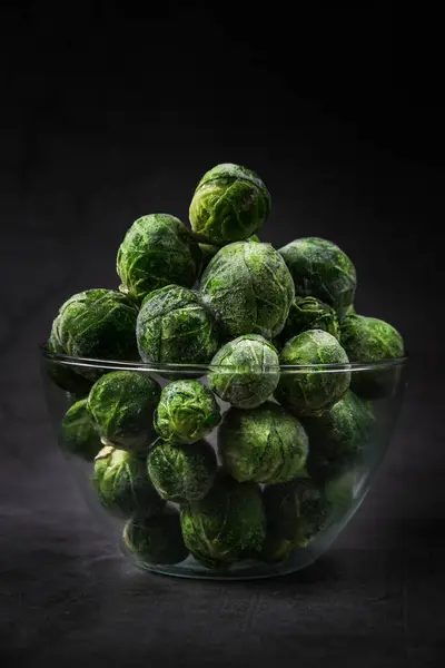 En glasskål med frysta Brysselkålhuvuden täckta med hesparfrost. Frysta gröna grönsaker. konservering av vintermat. Kallt från frysen. mörk bakgrund vertikal — Stockfoto