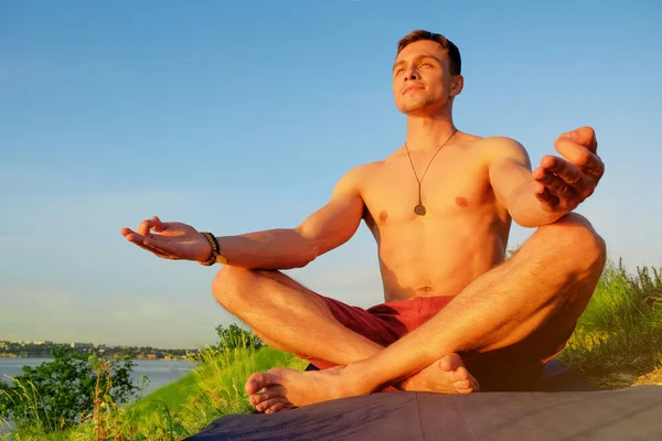 Ajuste hombre musculoso haciendo yoga junto al agua sentado en una pose de loto con las manos en om mudra. Naranja sol puesta del sol tranquilidad en el lago. El yogui masculino definió músculos. salud mental ejercicio tranquilo meditación — Foto de Stock
