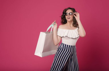 Güneş gözlüğü uzun dalgalı saçlı Genç bir kız onu gözler ve ağız sürpriz geniş açıldı. Onun elinde büyük bir paket için işaret eder. İzole pembe arka plan metin için yer ile üzerinde duran