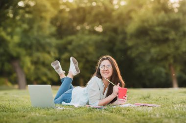 Genç bir öğrenci üniversitenin önünde yeşil çim üzerinde yatıyor ve final sınavları için hazırlanıyor. O bilimsel makaleler internette tarar ve yararlı notlar not defterinde yapar