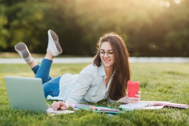 Genç bir öğrenci üniversitenin önünde yeşil çim üzerinde yatıyor ve final sınavları için hazırlanıyor. O bilimsel makaleler internette tarar ve yararlı notlar not defterinde yapar