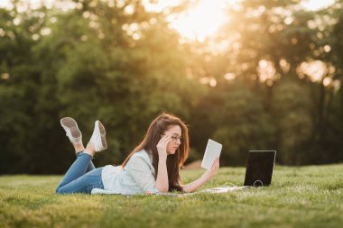 Genç bir öğrenci üniversitenin önünde yeşil çim üzerinde yatıyor ve final sınavları için hazırlanıyor. O bilimsel makaleler internette tarar ve yararlı notlar not defterinde yapar