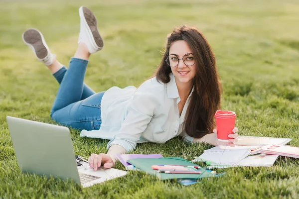 Genç bir öğrenci üniversitenin önünde yeşil çim üzerinde yatıyor ve final sınavları için hazırlanıyor. O bilimsel makaleler internette tarar ve yararlı notlar not defterinde yapar