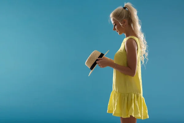 短い黄色い夏のドレスの若いブロンドの女の子は青い背景の上に立って 麦藁帽子を保持しています 少女は それを買うかと考えています — ストック写真